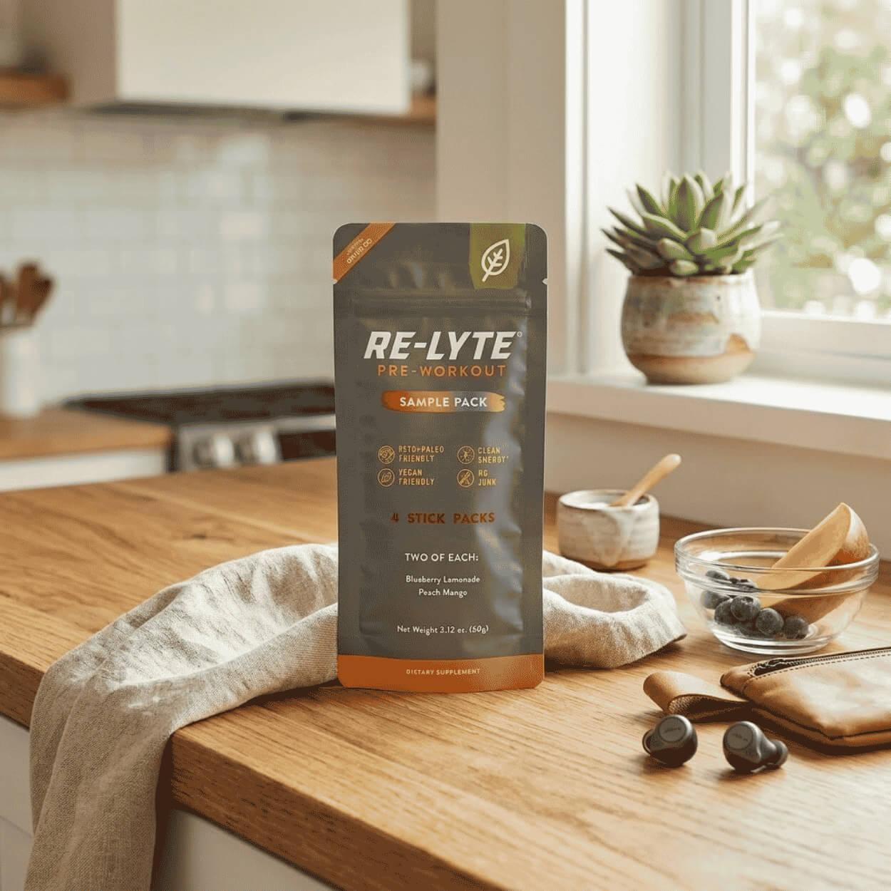 Na dřevěné kuchyňské lince stojí šedý sáček doplňku stravy Re-Lyte Pre-Workout vedle lněné utěrky, misky s ovocem, bezdrátových sluchátek a rostliny v květináči.